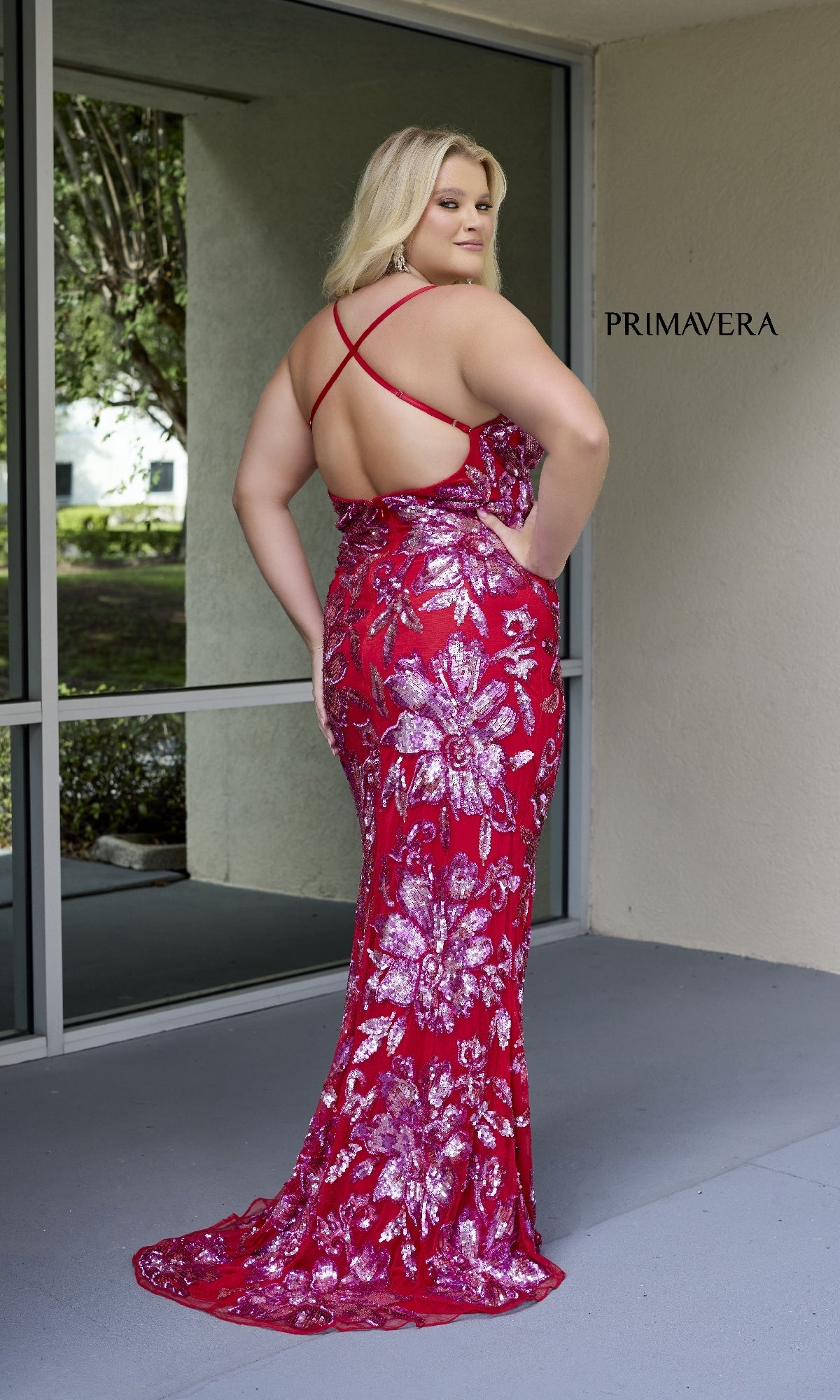A woman models the Primavera 14076, a long plus-size prom dress with floral sequins, low back, and crisscross straps, posing near a window.